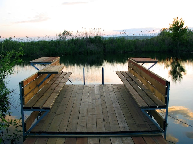 Place to rest on small river, wooden benches, pier, sunset, green plants, clouds are reflected