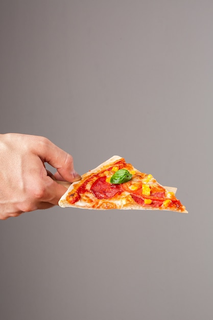 Plak van heerlijke hete eigengemaakte pizza in hand op grijze achtergrond. pizza - verse zelfgemaakte pizza met peperoni, kaas en tomatensaus, tomaten, suikermaïs en basilicum met kopie ruimte.