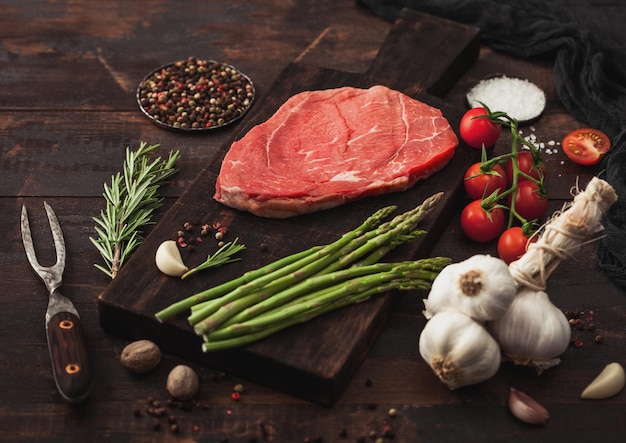 Plakje rauw vers rundvlees smoren steak op snijplank met knoflook asperges en tomaten met zout en peper met rozemarijn en vleesvork op houten achtergrond