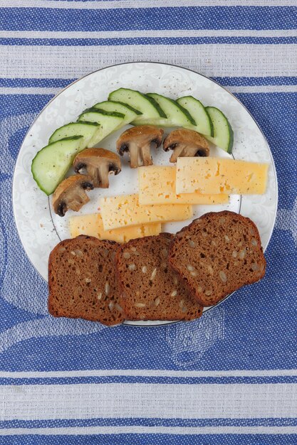 Plakjes gele harde kaas met plakjes komkommer plakjes gebakken champignons en graanbrood op witte plaat op een blauw servet Close-up van een gezonde snack Gezond voedselconcept