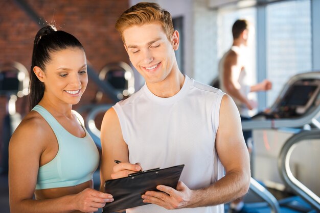 Foto pianificare il suo tempo in palestra. bel giovane istruttore maschio in piedi vicino alla bella donna e mostrando qualcosa nei suoi appunti mentre le persone corrono sul tapis roulant in background