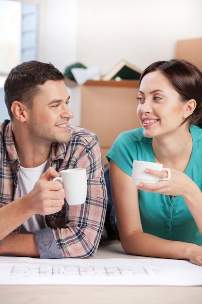 Planning a house improvement. Cheerful young couple lying on the floor and drinking coffee while cardboard boxes laying on the background