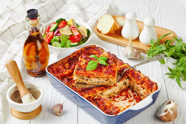 Plantaardige lasagne met tofu, champignons, passatasaus in een ovenschaal geserveerd met een frisse salade van sla, tomaat, komkommer, rode ui en basilicumblaadjes op een witte houten ondergrond, close-up