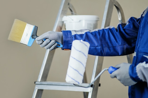 Il pittore di gesso è pronto per dipingere il muro. nelle mani di un rullo e pennello. una scala a pioli e un secchio di vernice sullo sfondo.