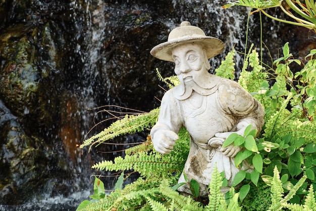 Plaster statue of a man with a beard in a hat among the fern.