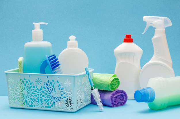 Plastic basket, bottles of dishwashing liquid, glass and tile cleaner, brushes, garbage bags on blue background. Washing and cleaning concept.