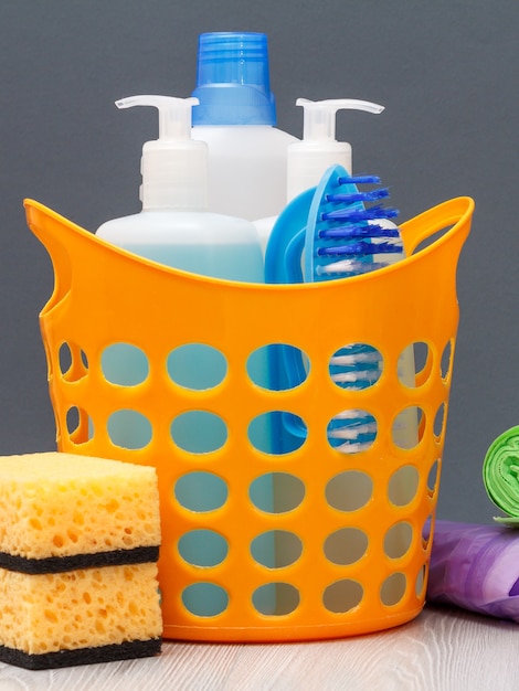 Plastic basket with bottles of dishwashing liquid, tile cleaner, detergent for microwave ovens and stoves, garbage bags and sponges on wooden and gray background. Washing and cleaning concept.