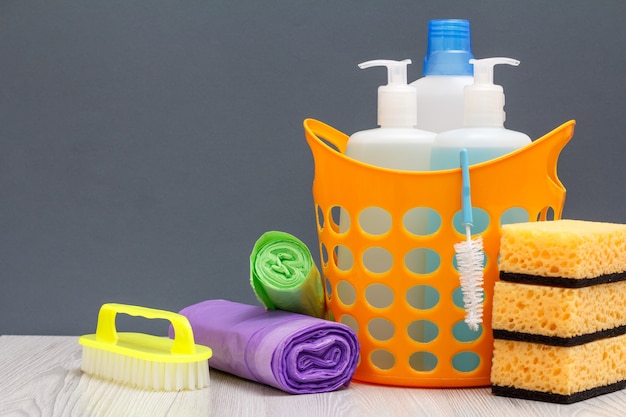 Plastic basket with bottles of dishwashing liquid, tile cleaner, detergent for microwave ovens and stoves. Sponges, garbage bags and brush on wooden and gray background. Washing and cleaning concept.