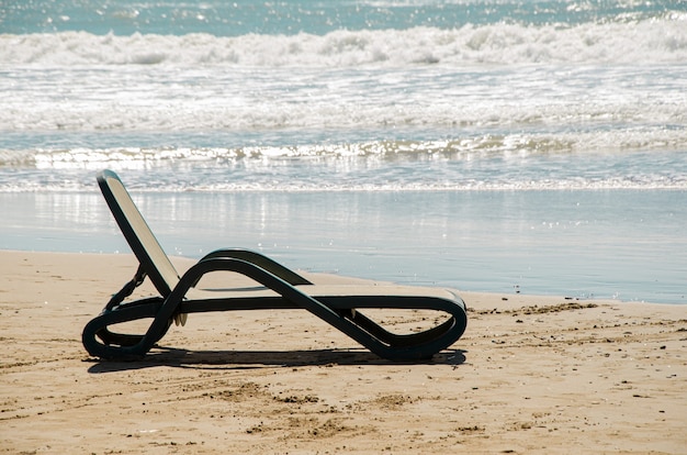 Plastic beach lounger stands on sandy beach at waters edge against backdrop of sea waves.