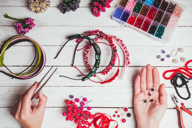 Plastic berries, flowers, beads and instruments for doing handmade headbands. Top view with hands