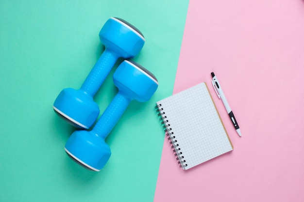 Plastic blue dumbbells, notepad on  pastel background