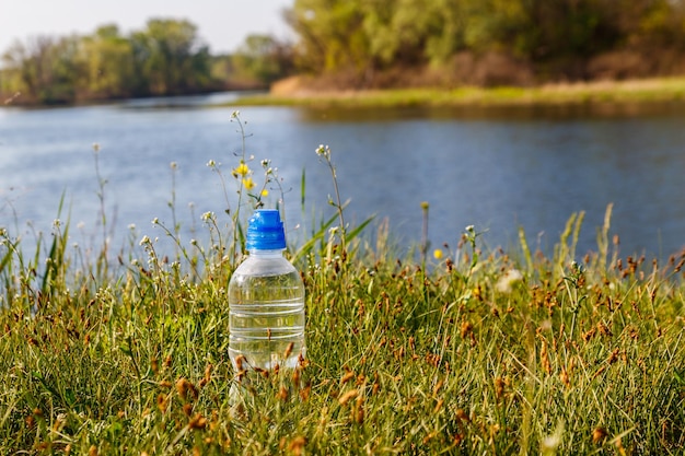 Пластиковая бутылка со свежей питьевой водой в зеленой траве на берегу реки