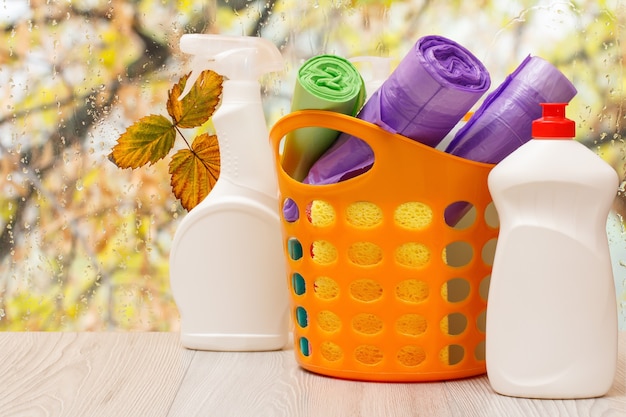 Plastic bottles of dishwashing liquid, basket with garbage bags, sponges in front of the window with water drops and autumn leaves. Washing and cleaning concept.