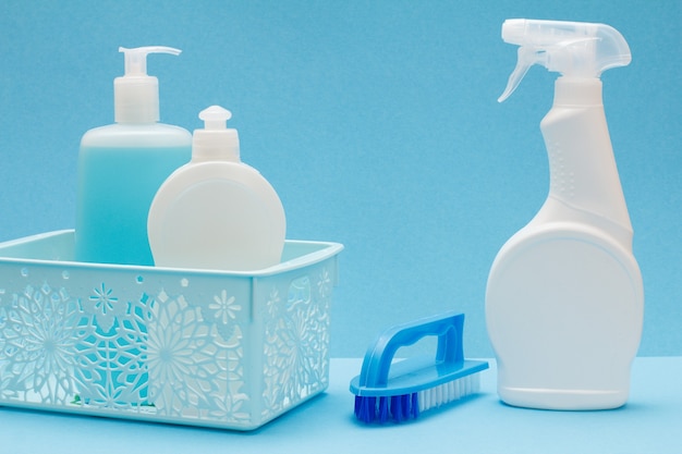 Plastic bottles of dishwashing liquid, glass and tile cleaner in basket and brush on blue background. Washing and cleaning concept.