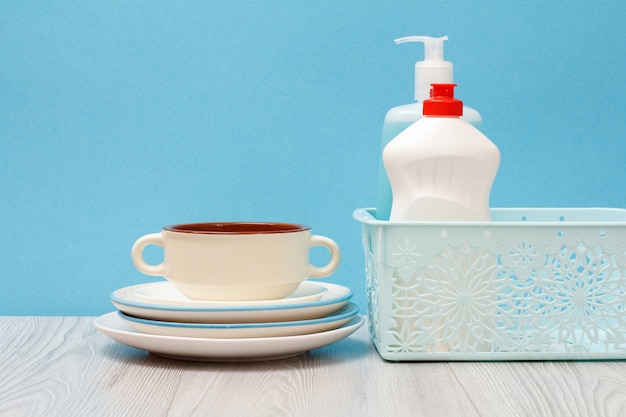 Пластиковые бутылки жидкости для мытья посуды, очиститель стекла и плитки в корзине, чистые тарелки и миску на синем фоне. Концепция стирки и очистки.