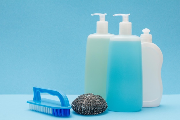 Plastic bottles of dishwashing liquid, glass and tile cleaner, brush and metal sponge on blue background. Washing and cleaning concept.