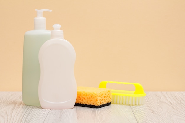 Plastic bottles of dishwashing liquid, glass and tile cleaner, brush and sponge on wooden boards and beige background. Washing and cleaning concept.