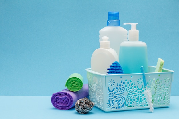 Plastic bottles of dishwashing liquid, glass and tile cleaner, brushes in basket, sponge, garbage bags on blue background. Washing and cleaning set.