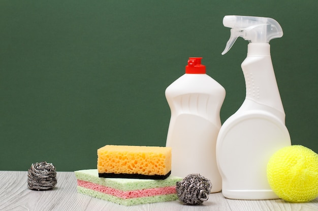 Plastic bottles of dishwashing liquid, glass and tile cleaner and sponges on green background. Washing and cleaning concept.