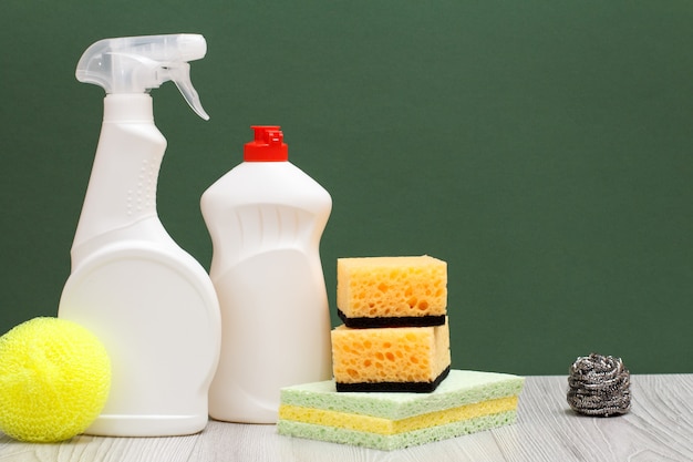 Plastic bottles of dishwashing liquid, glass and tile cleaner and sponges on laminate flooring and green background. Washing and cleaning concept.