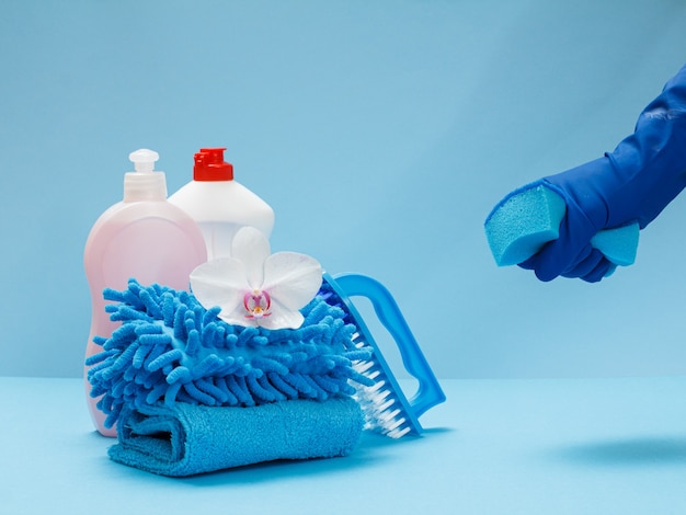 Plastic bottles of dishwashing liquid, rags, a brush, a white orchid flower and a hand in a rubber glove holding a sponge on the blue background. Washing and cleaning set.