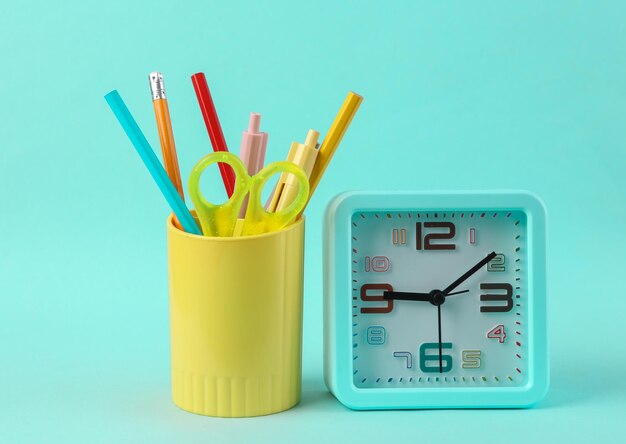 Plastic cup with school stationery drawing tools and alarm clock on blue background