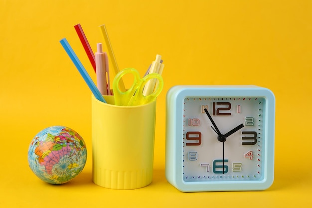 Plastic cup with school stationery drawing tools and alarm clock globe on yellow background back to school