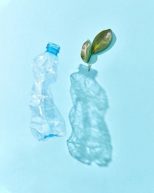 Plastic drijvende fles met schaduwen en groen plantentakje op een pastelblauwe muur, exemplaarruimte. Milieuvervuiling van plastic.