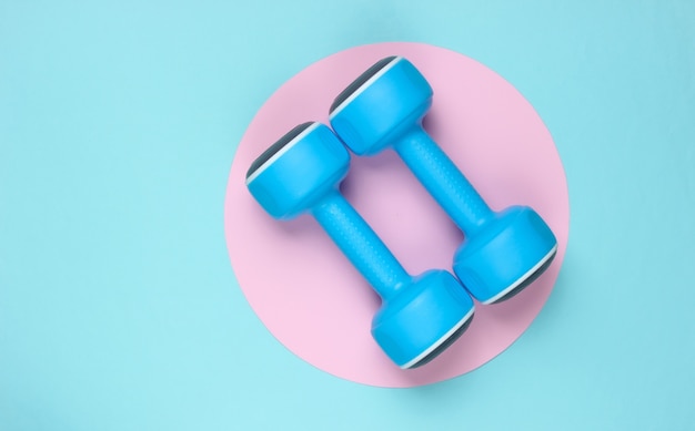 Plastic dumbbell on blue background with pink pastel circle. Top view. Minimalistic sport concept. Top view