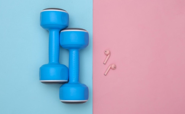 Plastic dumbbells and wireless headphones on a blue-pink pastel background. Top view