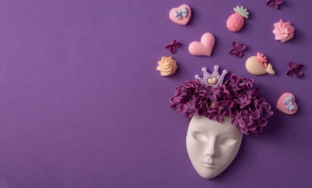 Plastic face mask decorated with lilac flowers and miniatures of crown and sweets on purple background