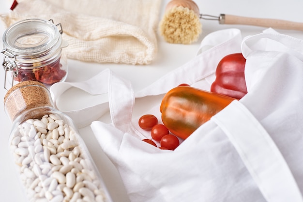 Plastic free reusable products, zero waste concept. Fresh vegetables, beans in a glass bottle and textile shopping bag on white background