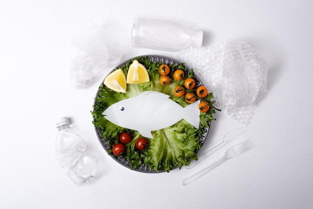 Plastic garbage white plastic fish with vegetables on plate on\
white background concept of water and fish pollution unhealthy\
food