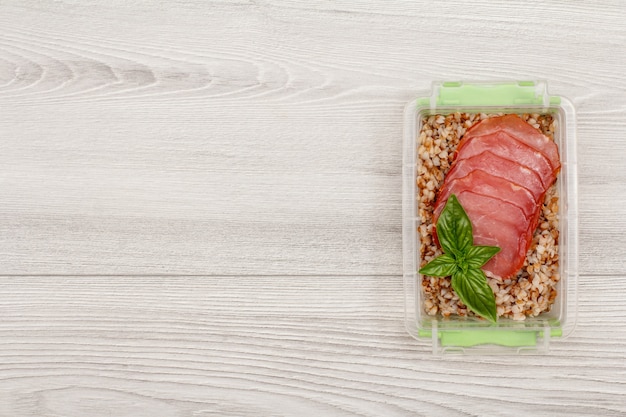 Plastic meal prep container with boiled buckwheat porridge and slices of meat on grey wooden background. Top view with copy space