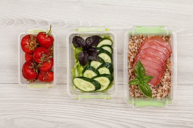 Plastic meal prep containers with fresh strawberries, cucumbers and salad, boiled buckwheat porridge and slices of meat on grey wooden background. Top view