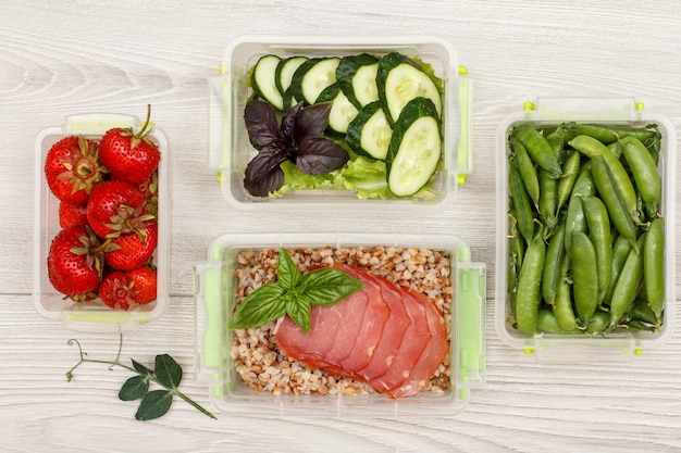 Plastic meal prep containers with fresh strawberries, green peas, cucumbers and salad, boiled buckwheat porridge and slices of meat on grey background. Top view