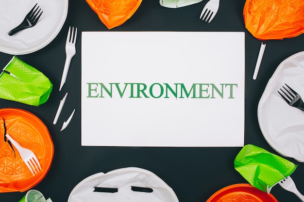 Plastic pollution and environment protection. Paper with word Environment in the centre of disposable colorful broken plastic plates and forks on dark surface.