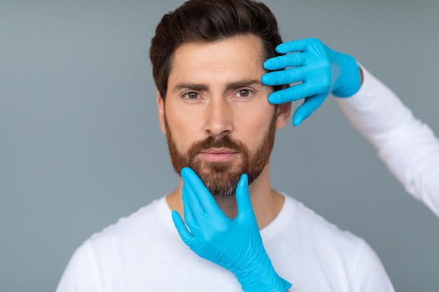 Концепция пластической хирургии и эстетической косметологии Руки косметолога в защитных медицинских перчатках касаются мужского лица