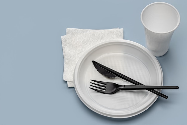 Plastic utensils. disposable white plates, cup and black knife,\
fork on a gray background.