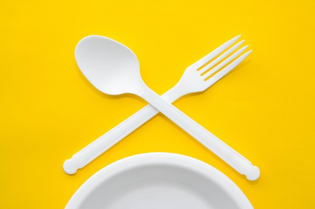 Plastic white crossed fork, spoon and plate on yellow background. Cooking utensil. Cutlery sign. Top view. Minimalist Style. Copy, empty space for text