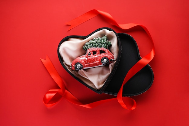 Foto plat lag kerst samenstelling. kleine rode speelgoedauto in een zwarte doos in de vorm van een hart met een rood lint op rood.