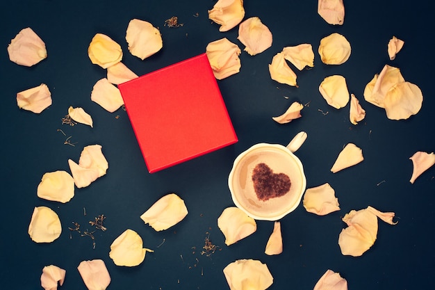 Plat leggen. Rode doos, een koffiemok met room en kaneel in de vorm van een hart met roze bloemblaadjes op een donkerblauwe achtergrond. Kopieer ruimte op de doos, voor het merk. Geschikt voor indeling. vignettering.