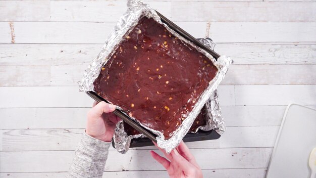 Plat leggen. Stap voor stap. Gesmolten chocolade scheppen in bakpannen bekleed met kookfolie om macadamia-nootfudge te maken.