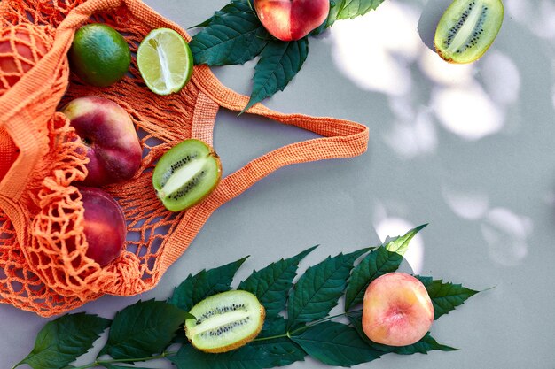 Plat leggen van eco-vriendelijke mesh boodschappentas met fruit perzik, kiwi, limoen op grijs oppervlak in zonlicht, zomertijd. Kruidenier concept, kopieer ruimte, bovenaanzicht.