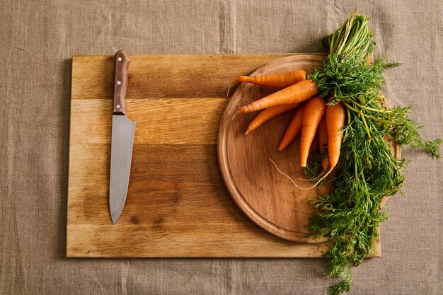 Plat leggen van een grote bos verse wortelen met groene toppen en een keukenmes op een houten snijplank op een linnen tafelkleed. Voedselachtergrond van seizoensgebonden rauwe groenten