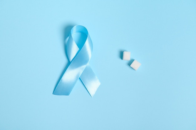 Plat leggen van pure geraffineerde witte suikerklontjes en blauw satijnen lint, symbool van Werelddiabetesbewustzijnsdag 14 november. Geïsoleerd over gekleurde blauwe achtergrond, kopieer ruimte voor medische reclame.