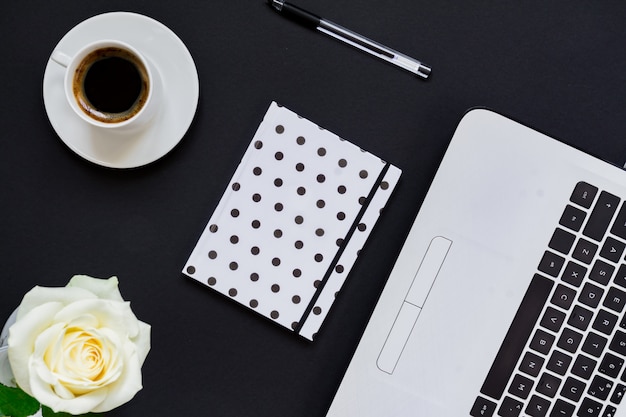 Plat liggende, bovenaanzicht kantoor tafel bureau. Werkruimte met laptop, witte roos, polka dot agenda en koffiemok op zwart.