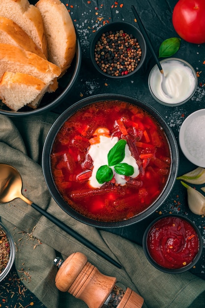 A plate of beetroot soup with sour cream and rosemary, bread and tomato sauce on a black concrete background. Top view, vertical.