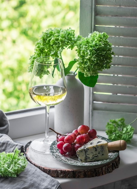 Plate of cheese grapes glass of white wine on the windowsill with wooden shutters window with a view of the summer garden Rustic still life