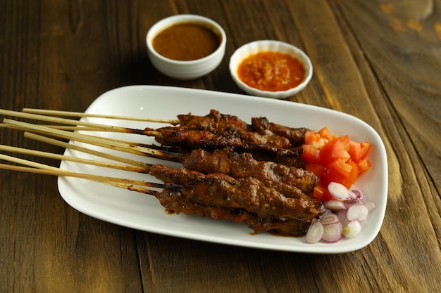 A plate of Chicken Satay served on a white plate with sliced onions tomatoes and a bowl sambal